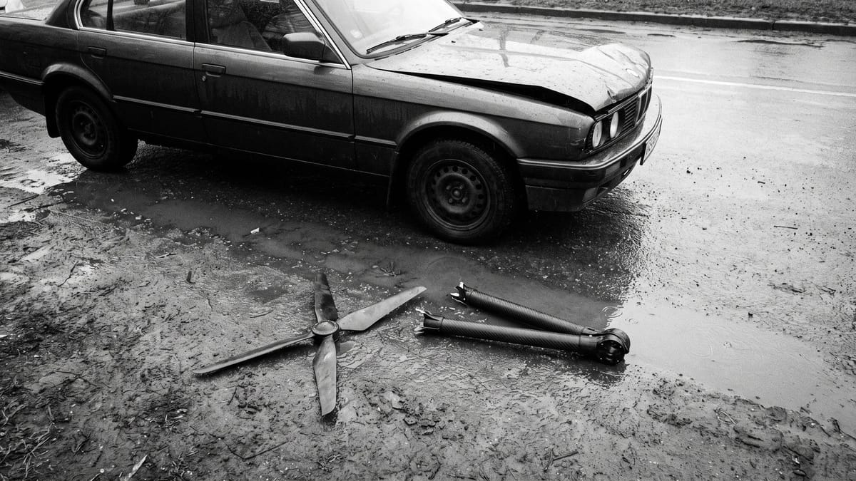 Elica spezzata e braccio di un drone sull'asfalto bagnato accanto al cofano ammaccato di un'auto, in bianco e nero