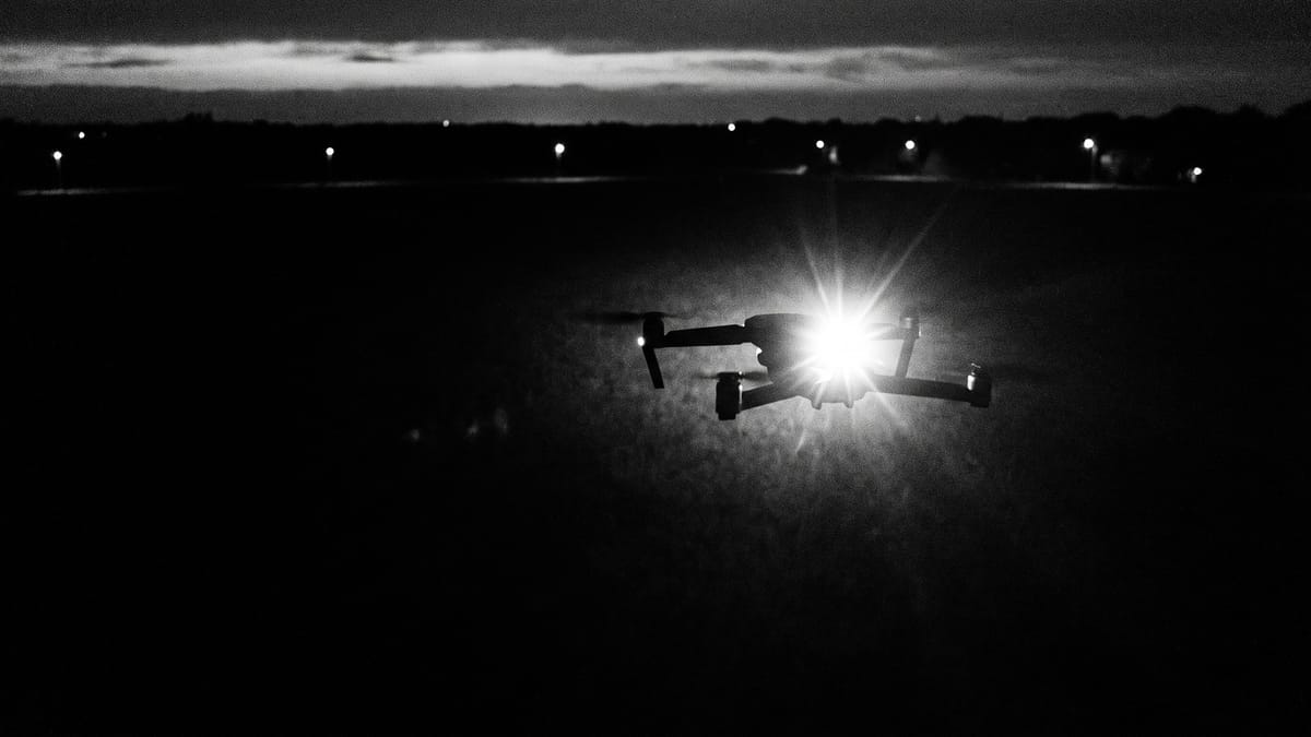 Campo notturno con un drone in volo basso e una luce accesa a bordo che risalta nel buio, fotografia in bianco e nero