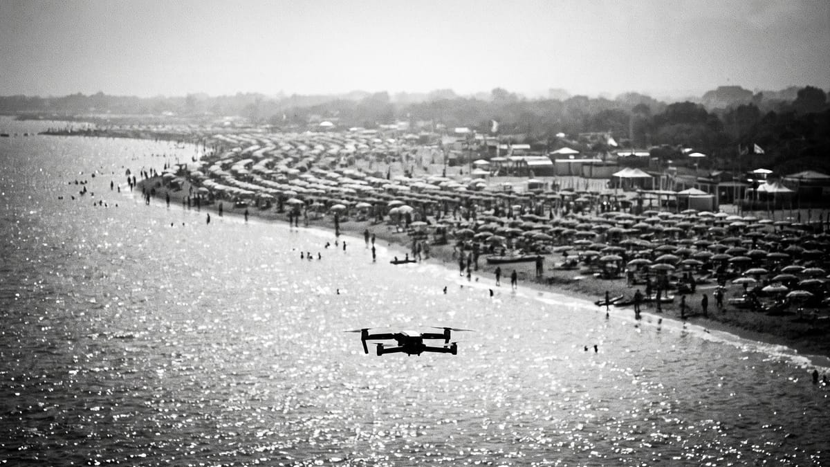 Spiaggia affollata vista da lontano con file di ombrelloni e un piccolo drone in volo sull'acqua in primo piano, bianco e nero